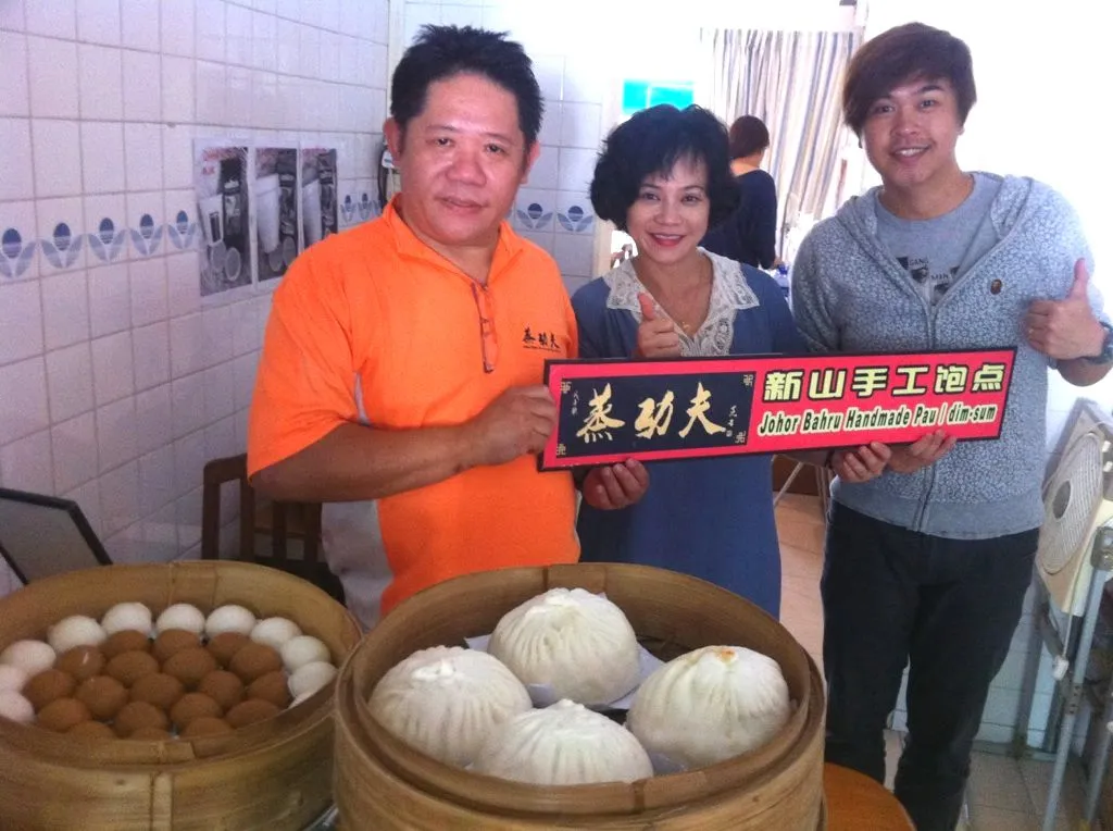 Three people behind baskets of pau, holding a Kung Fu JB Pau sign